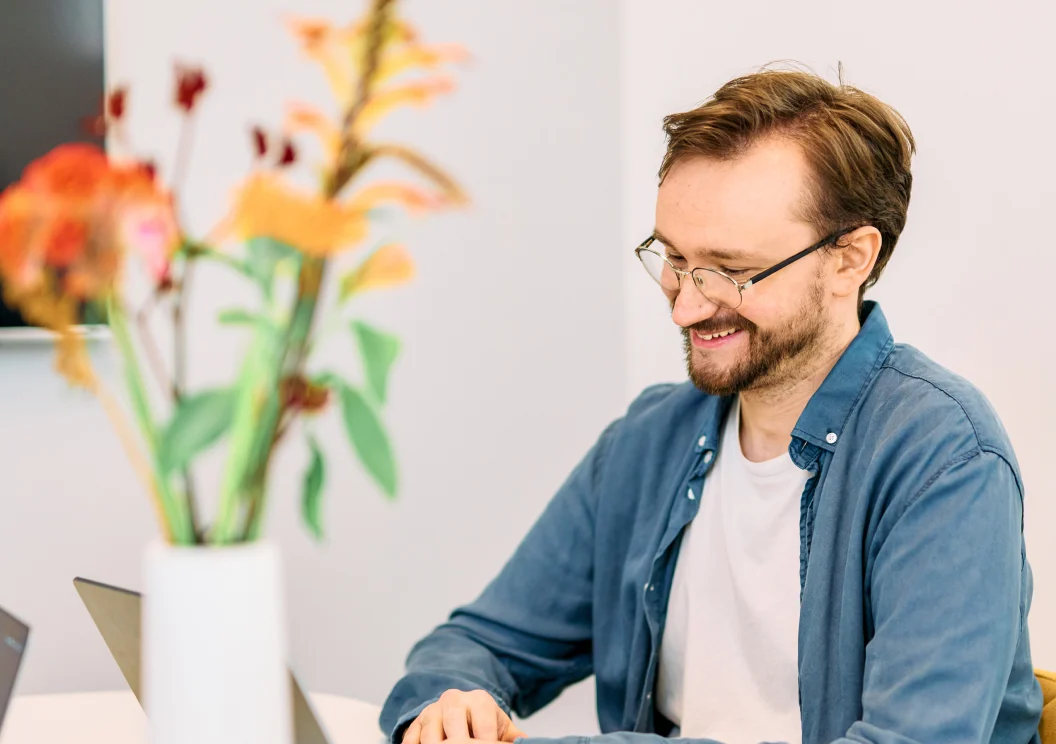 Man in a denim shirt smiling at his laptop with flowers in the foreground