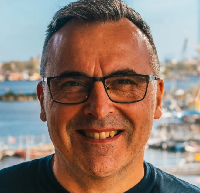 Portrait of Alexander smiling in glasses with a harbour skyline behind him