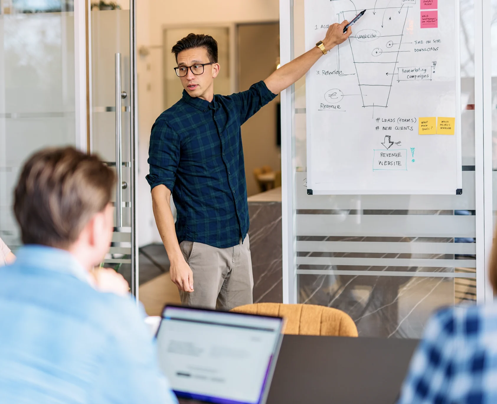 Man in green checkered shirt pointing at whiteboard with diagrams during office meeting.