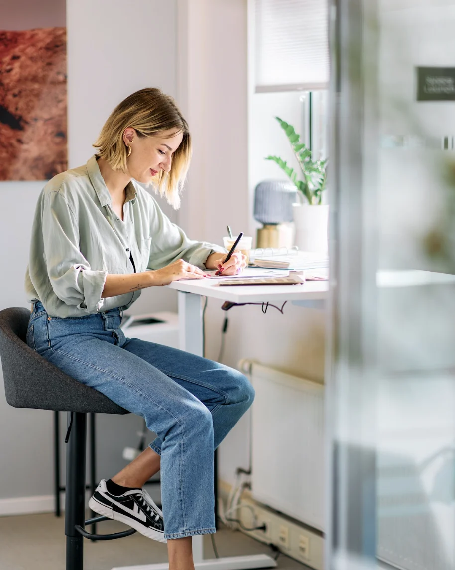 Woman in casual clothes sitting on a stool writing at a white desk with a plant nearby.