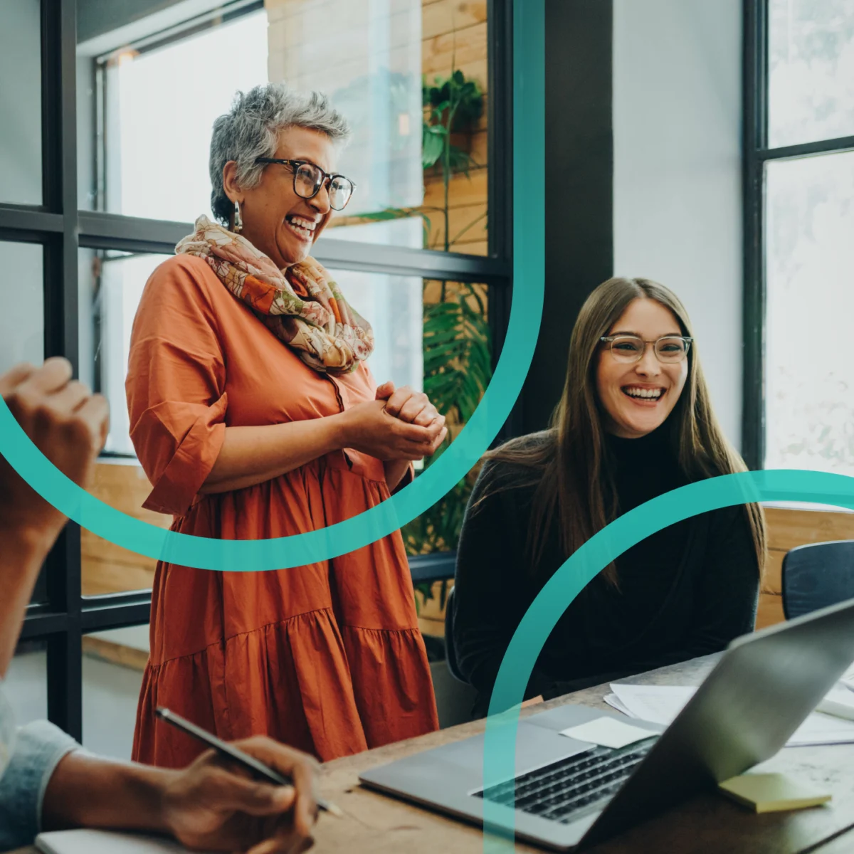 Two colleagues laughing together at a laptop in an office, framed by teal brand rings