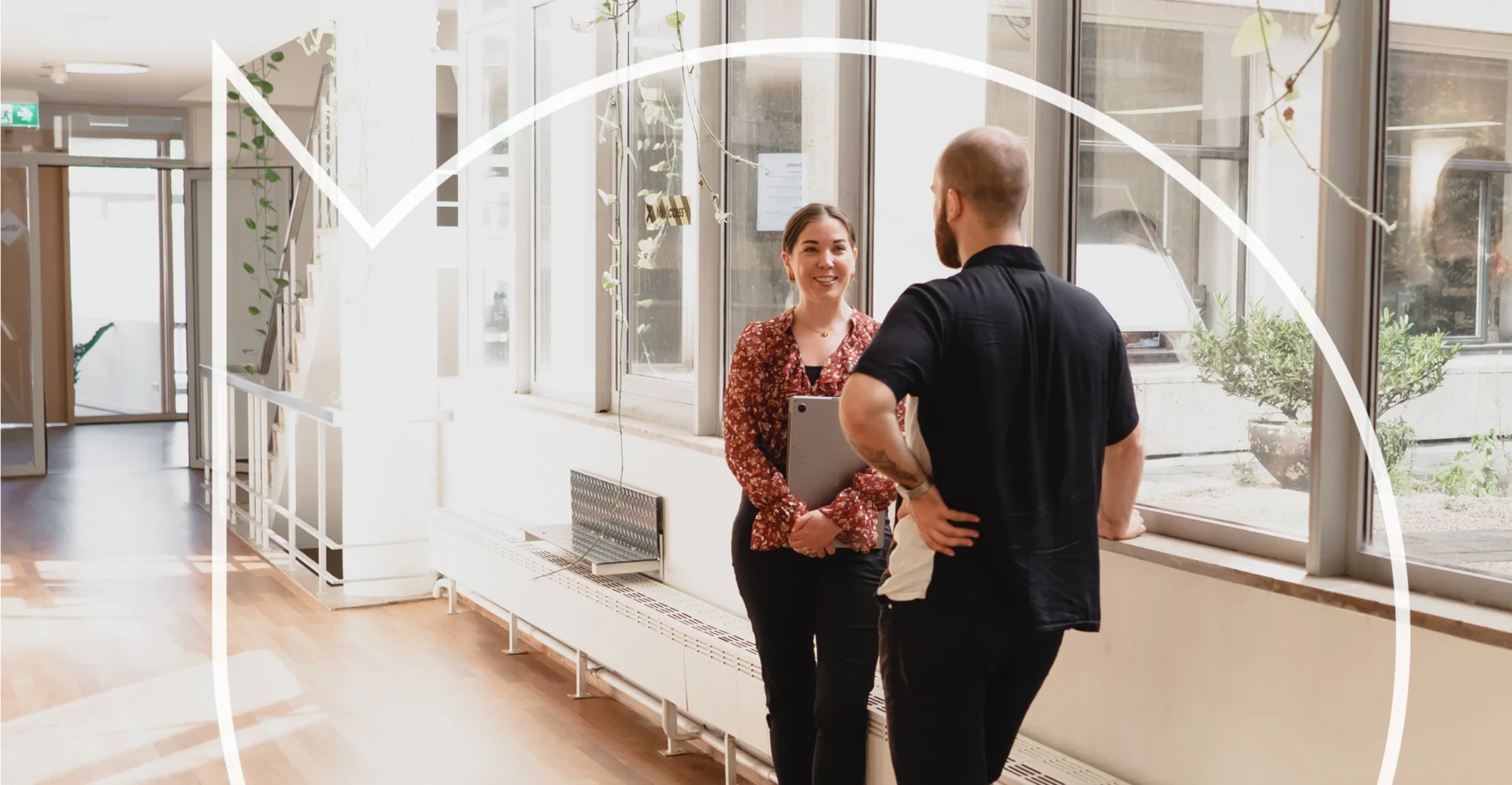 Two colleagues talking in a sunlit modern office hallway next to large windows.